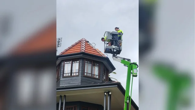 Two workers in an elevated work platform beside the red tiled turret roof of a dark timber home.