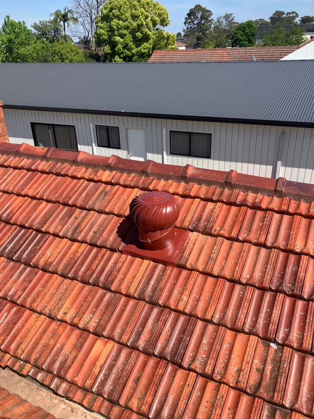 Red tiled roof with ridge tiles and a red roof ventilator, looking toward a grey metal roof.